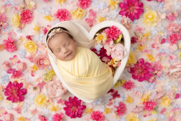 Sleeping newborn baby wrapped in yellow swaddle in a heart bowl with flowers all around in Chicago studio