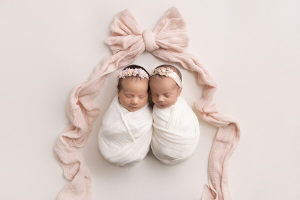 Sleeping newborn twin girls wrapped in neutral swaddle in Chicago studio with pink bow